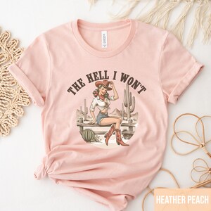 May include: A pink t-shirt with a graphic of a woman in a cowboy hat sitting on a fence with the text "The Hell I Won't".