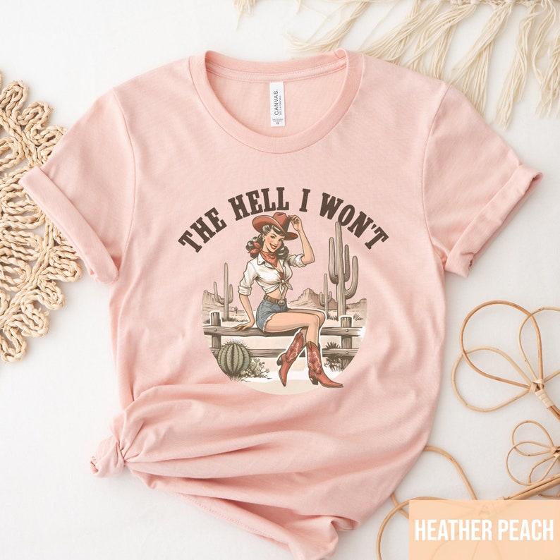 May include: A pink t-shirt with a graphic of a woman in a cowboy hat sitting on a fence with the text "The Hell I Won't".