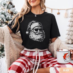 May include: Black t-shirt featuring a white graphic of Santa Claus wearing sunglasses. The Santa graphic has a beard and hat. The person in the image is wearing red and white plaid pajama pants and holding a mug with a Santa Claus design.