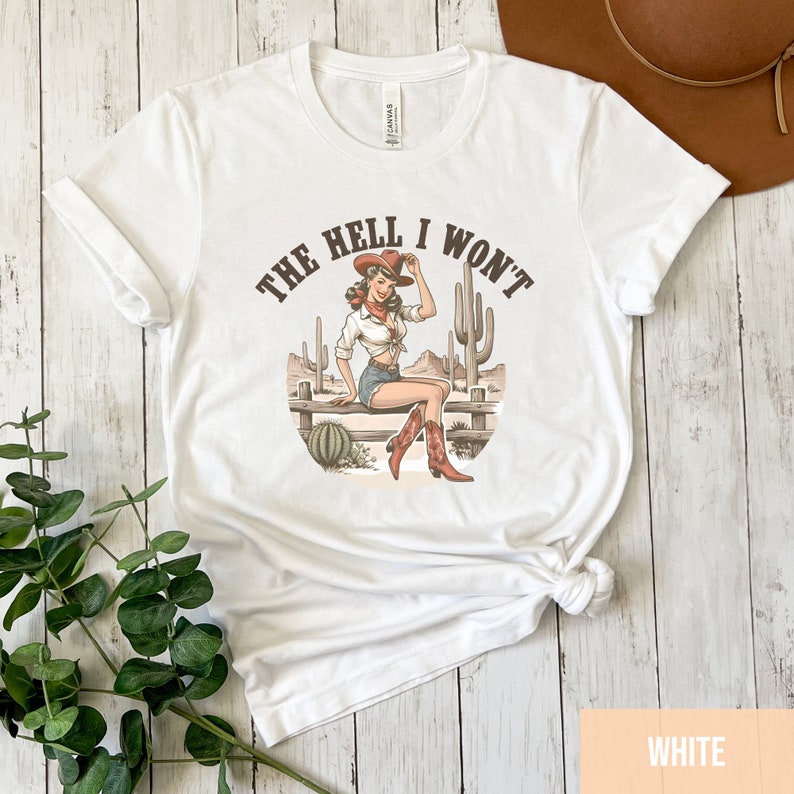 May include: White t-shirt with a graphic of a woman wearing a cowboy hat and boots, sitting on a fence with cacti in the background. The text "THE HELL I WON'T" is printed on the shirt.