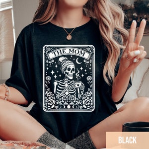 May include: Black t-shirt featuring a white graphic of a skeleton wearing a beanie and holding a cup of coffee. The graphic is surrounded by flowers and the text "The Mom" is above the skeleton. The text "Black" is visible in the bottom right corner of the image.