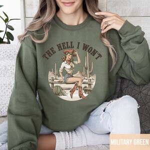 May include: A green sweatshirt with the text "The Hell I Won't" in a circle. Inside the circle is a cartoon image of a woman wearing a cowboy hat and boots, sitting on a fence with cacti in the background.