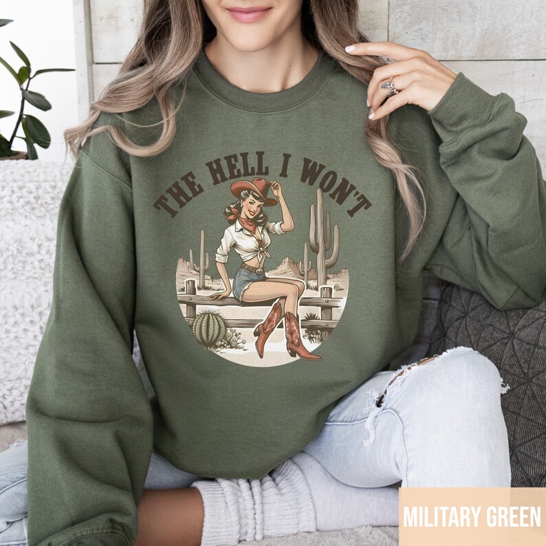 May include: A green sweatshirt with the text "The Hell I Won't" in a circle. Inside the circle is a cartoon image of a woman wearing a cowboy hat and boots, sitting on a fence with cacti in the background.