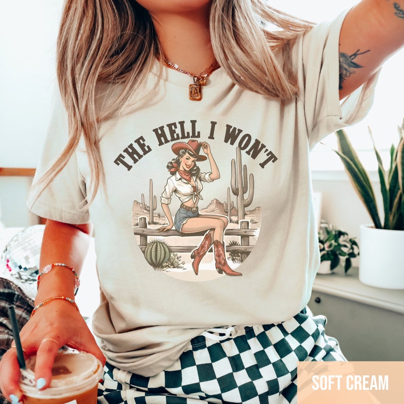 May include: A cream colored t-shirt with a vintage graphic of a woman in a cowboy hat sitting on a fence with the text "The Hell I Won't".