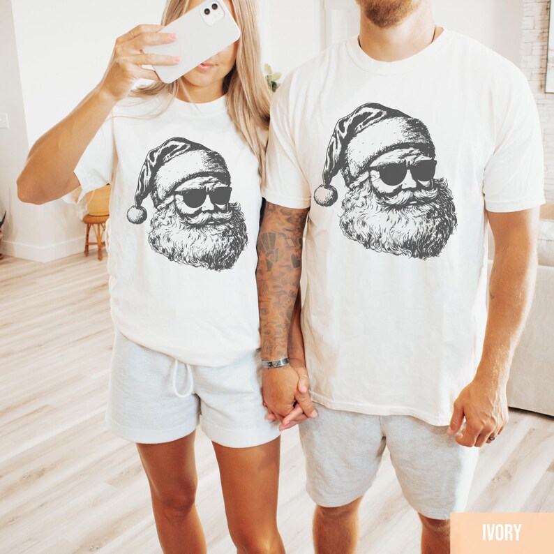 May include: Ivory-colored t-shirts with a black graphic of Santa Claus wearing sunglasses. The image shows a couple wearing matching shirts and gray shorts. The shirts have a crew neck and short sleeves. The Santa graphic includes a hat, beard, and mustache.