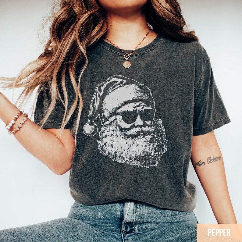 May include: Dark grey t-shirt featuring a black and white illustration of Santa Claus wearing sunglasses. The Santa graphic includes his hat, beard, and mustache. The shirt is a relaxed fit and has a vintage look.