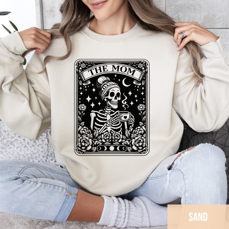 May include: A beige sweatshirt featuring a black and white graphic of a skeleton wearing a beanie and holding a cup of coffee. The graphic is in the style of a tarot card and says "The Mom" at the top.
