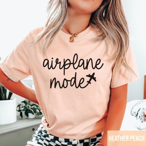 Pu&ograve; includere: Una maglietta rosa chiaro con il testo "airplane mode" in scrittura nera con una piccola icona di aereo accanto alla parola "mode". La maglietta &egrave; indossata da una persona con lunghi capelli biondi. Il testo "HEATHER PEACH" &egrave; visibile nell'angolo in basso a destra dell'immagine.