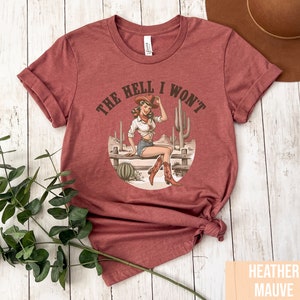 May include: A heather mauve t-shirt with a vintage illustration of a woman wearing a cowboy hat and boots. The text "The Hell I Won't" is printed on the shirt.