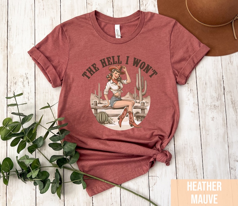 May include: A heather mauve t-shirt with a vintage illustration of a woman wearing a cowboy hat and boots. The text "The Hell I Won't" is printed on the shirt.