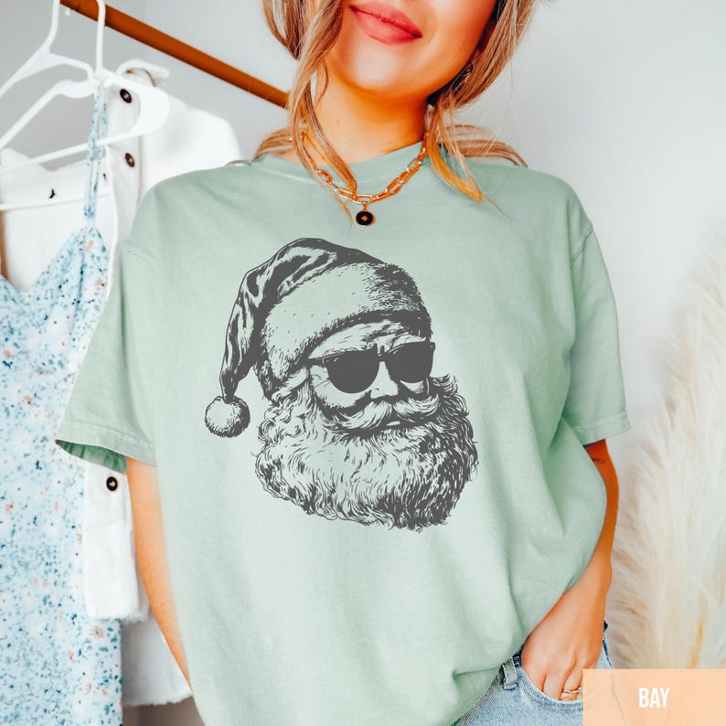 May include: A light green t-shirt featuring a black and white graphic of Santa Claus wearing sunglasses. The Santa illustration has a long beard and is wearing a Santa hat. The shirt is a casual style.