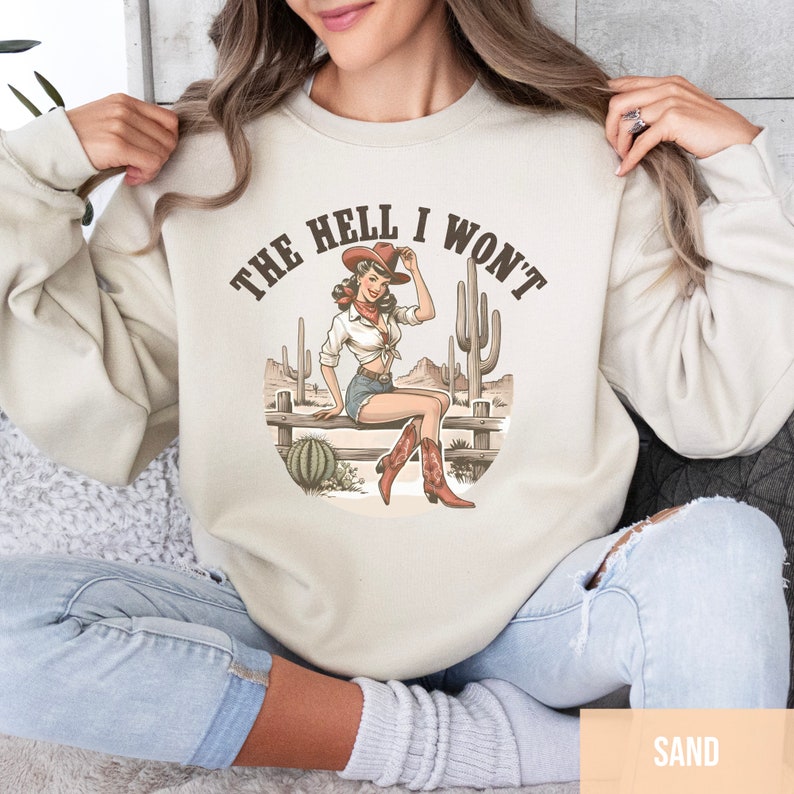 May include: A beige sweatshirt with a graphic print of a woman in a cowboy hat and boots sitting on a fence with cacti in the background. The text "THE HELL I WON'T" is printed above the graphic.