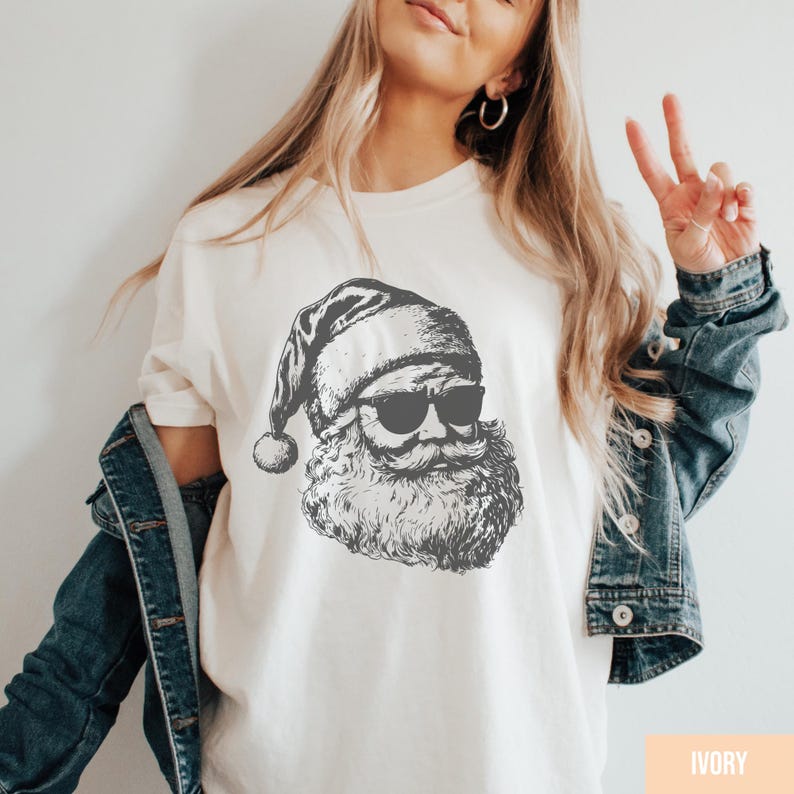 May include: Ivory-colored t-shirt featuring a black and white illustration of Santa Claus wearing sunglasses. The Santa graphic includes his hat, beard, and mustache. The shirt is a casual style, suitable for the holiday season.