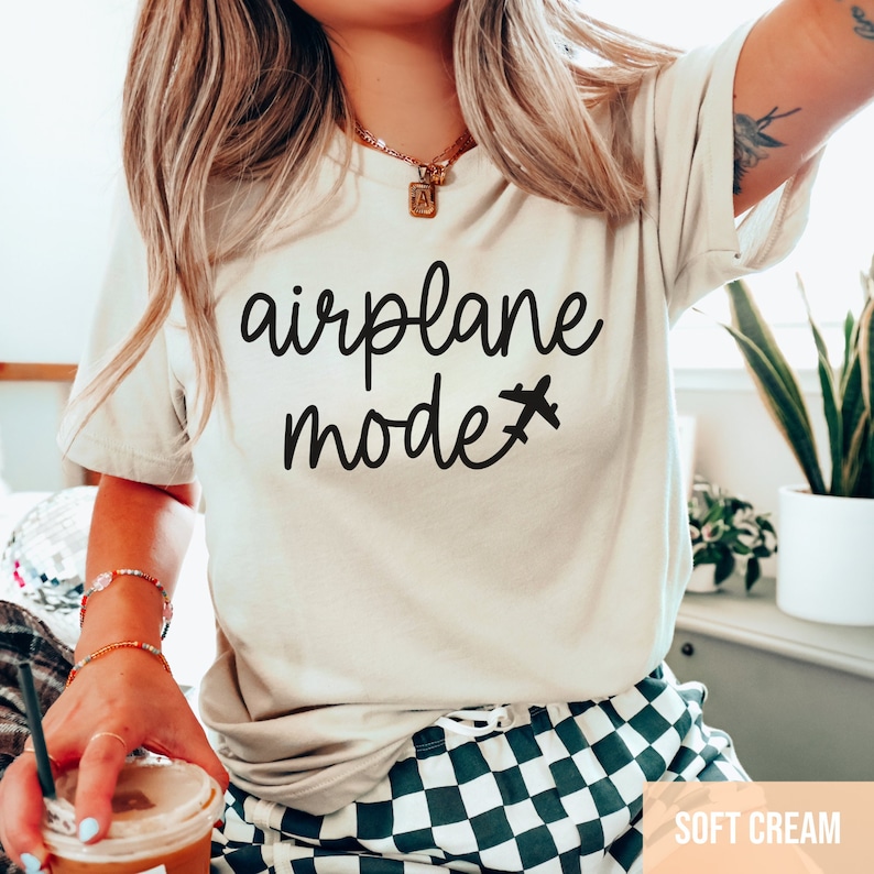 Pu&ograve; includere: Una maglietta bianca con la scritta "airplane mode" e una piccola icona di aereo in nero. La maglietta &egrave; indossata da una persona con i capelli lunghi e biondi. Lo sfondo &egrave; un'immagine sfocata di un motivo a scacchi e una pianta.