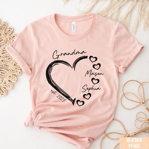 May include: A light pink t-shirt with a black heart design. The heart contains the word "Grandma" and the names "Mason" and "Sophia". The text "est. 2023" is also included. The shirt is made of soft material.
