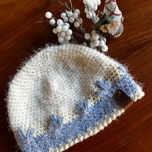 Può includere: Un cappello all'uncinetto bianco e blu con una texture soffice. Il cappello ha un'etichetta in pelle marrone con il testo "Betsy Boo's Hats".
