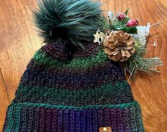 May include: A handmade beanie hat with a dark green faux fur pom-pom. The hat features a ribbed brim in shades of purple and blue, transitioning to green and purple stripes. A small tag reads "Beanie Boop Hats". Decorative pinecone and greenery are on the side.