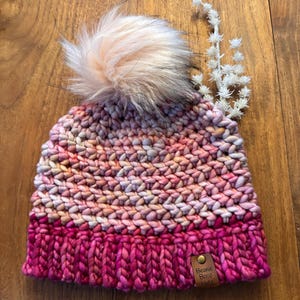 Może przedstawiać: Ręcznie robiona czapka beanie w odcieniach różu i kremu, zwieńczona puszystym, jasnoróżowym pomponem. Czapka ma głęboki, karmazynowy ściągacz i małą skórzaną metkę z napisem "Beanie Boop Hats".