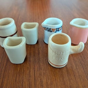 May include: A collection of small, ceramic mugs and cups in various shapes and colors. One mug has a handle and the words "THUMB FUN" printed on it. Other cups are white, beige, and pink, with decorative details.
