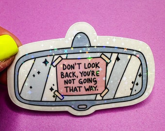 May include: A white sticker with a holographic finish depicting a rearview mirror with a pink note that reads "Don't look back, you're not going that way."