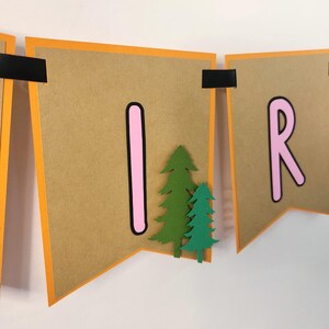 May include: A banner with brown pennants, orange borders, and black accents. The letters "I" and "R" are in pink, and green paper trees are attached to the pennants. The banner is likely for a birthday or party.