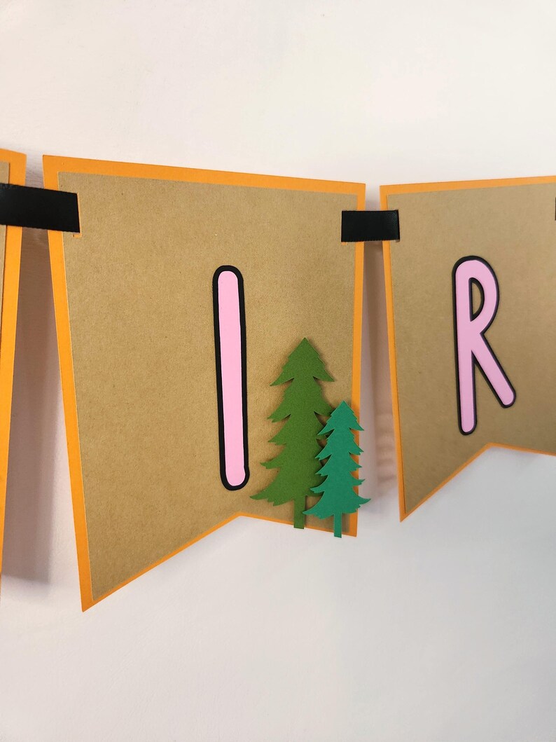 May include: A banner with brown pennants, orange borders, and black accents. The letters "I" and "R" are in pink, and green paper trees are attached to the pennants. The banner is likely for a birthday or party.