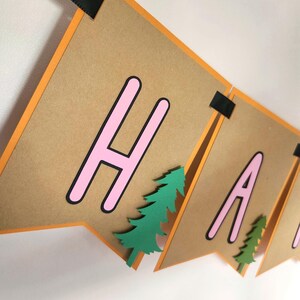 May include: A banner with brown pennants, orange borders, and black ribbon. The banner spells out "HAPPY" in pink letters with black outlines. Green paper trees are attached to the pennants, creating a festive decoration.