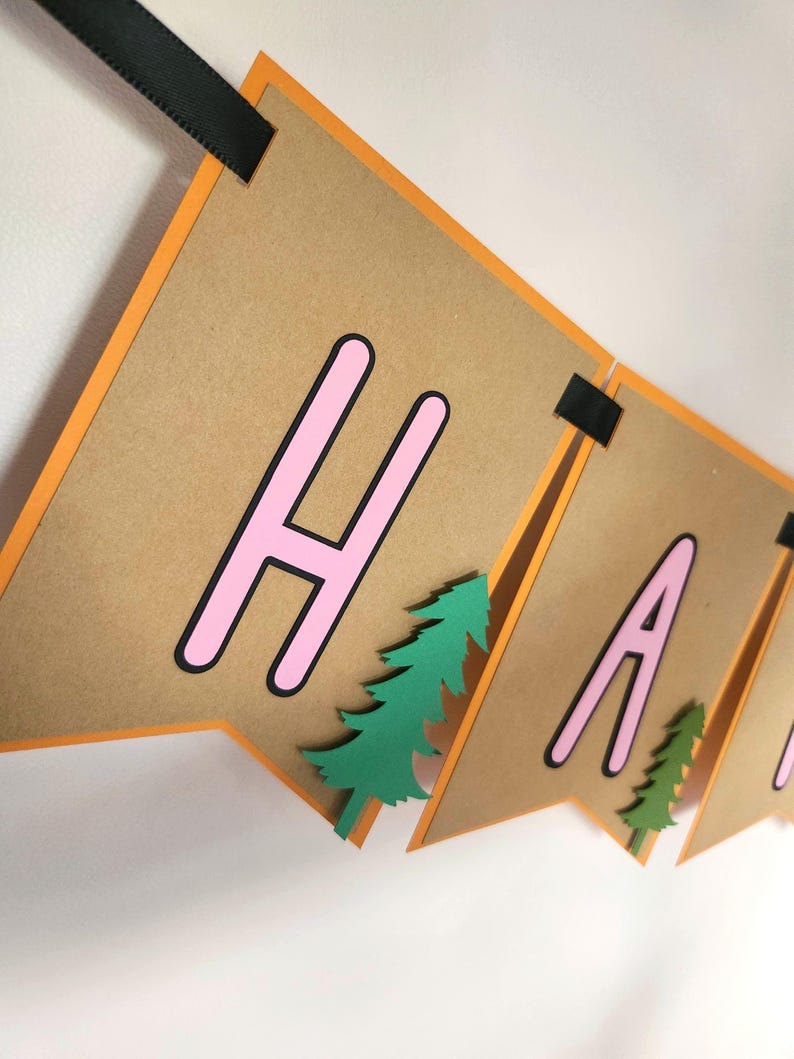 May include: A banner with brown pennants, orange borders, and black ribbon. The banner spells out "HAPPY" in pink letters with black outlines. Green paper trees are attached to the pennants, creating a festive decoration.