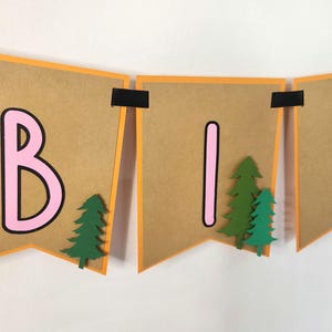 May include: A banner spelling out "BIR" in pink letters with black outlines on brown pennants. Each pennant has an orange border and is decorated with green paper trees. The banner is held together with black tape.