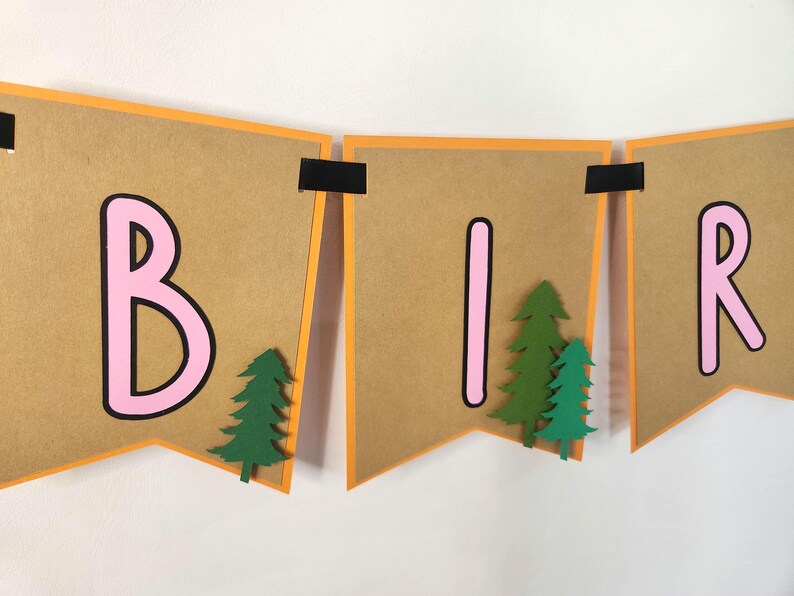 May include: A banner spelling out "BIR" in pink letters with black outlines on brown pennants. Each pennant has an orange border and is decorated with green paper trees. The banner is held together with black tape.