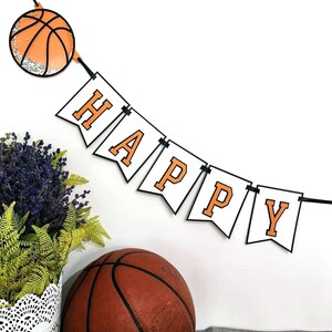 May include: A white banner with black trim and orange letters spelling "HAPPY" hangs from a black string. A basketball with orange and black markings is attached to the string. A second basketball is in the foreground.