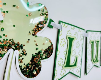 May include: A green and white banner with the word "LUCKY" in green letters. The banner has a shamrock with green and gold glitter inside.