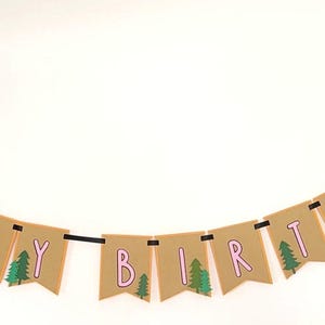 May include: A banner with the words "HAPPY BIRTHDAY" in pink letters on brown pennants. Green trees are printed on some pennants. The banner is strung on a black cord and has decorative elements of a campfire at each end.