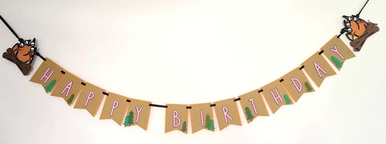 May include: A banner with the words "HAPPY BIRTHDAY" in pink letters on brown pennants. Green trees are printed on some pennants. The banner is strung on a black cord and has decorative elements of a campfire at each end.