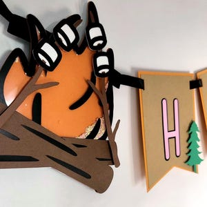 May include: A layered papercraft banner with a campfire and the word "HAPPY". The campfire features orange flames, brown logs, and white marshmallows. The banner flags are tan with pink letters and green trees.
