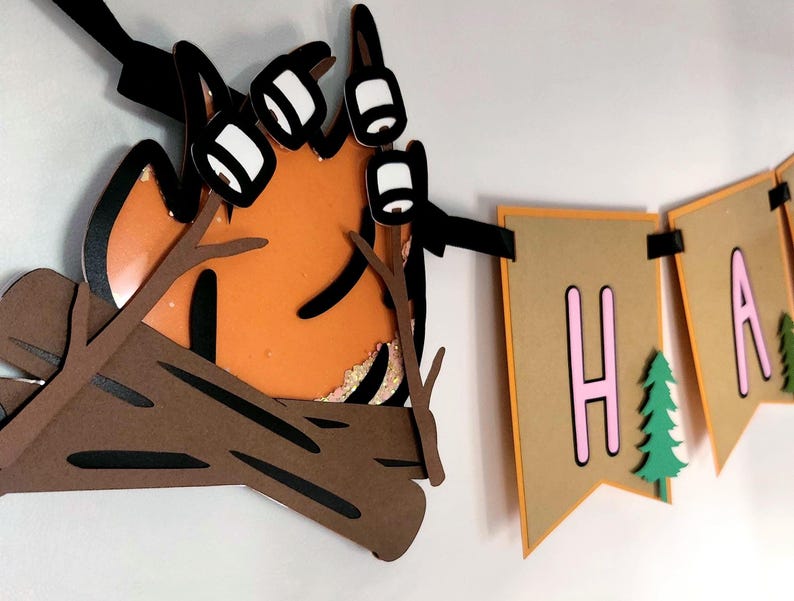 May include: A layered papercraft banner with a campfire and the word "HAPPY". The campfire features orange flames, brown logs, and white marshmallows. The banner flags are tan with pink letters and green trees.