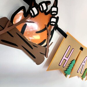 May include: A layered paper banner with the word "HAPPY" in pink letters, with green tree accents. A campfire with marshmallows on sticks and a log are also part of the design. The banner is brown and orange.