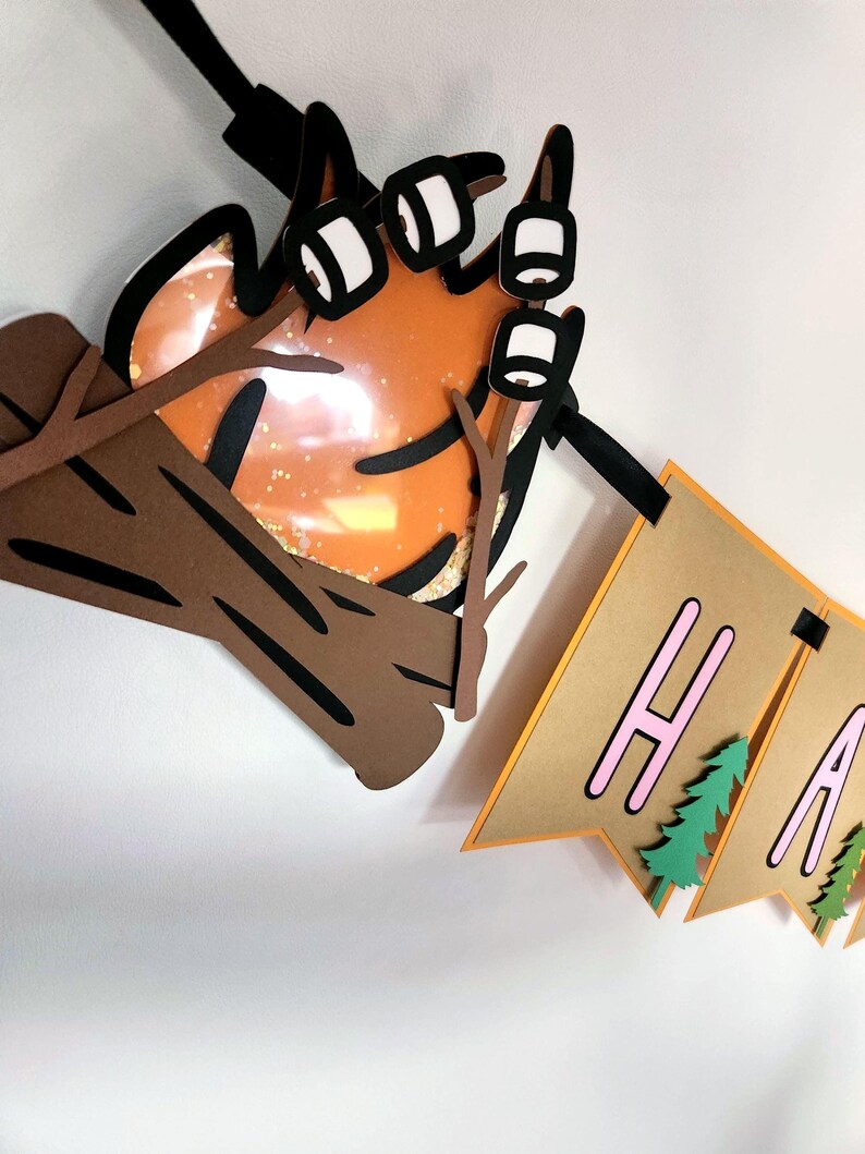 May include: A layered paper banner with the word "HAPPY" in pink letters, with green tree accents. A campfire with marshmallows on sticks and a log are also part of the design. The banner is brown and orange.