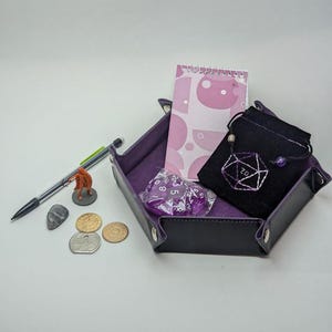 May include: Purple dice, a small purple pouch with a white "D20" embroidered on it, a small orange miniature figure, a purple dice tray, a notebook with a pink and white pattern, and a few coins on a white surface.