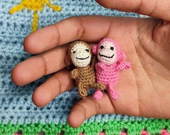 May include: Two small crocheted monkey toys, one brown and one pink, are held in a hand against a colorful crocheted background.