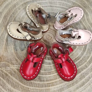 May include: Four pairs of miniature leather shoes. Two pairs are beige with white stitching, one pair is pink with white stitching, and one pair is red with white stitching.