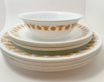 Puede incluir: Una pila de platos de cerámica blanca con un patrón floral dorado alrededor del borde. Un tazón de cerámica blanca con un patrón floral dorado alrededor del borde se sienta encima de los platos.