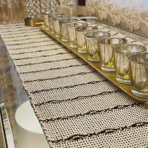 May include: A decorative table runner with a woven pattern of cream, black, and gold. A gold tray holds several small, clear, gold-toned glasses. The glasses are arranged in a row, suggesting a display for candles or small beverages.