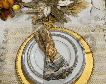 May include: Elegant table setting with gold chargers, white plates with silver trim, and patterned napkins. A gold poinsettia centerpiece and decorative pumpkins with candles add to the festive decor. Wine glasses complete the arrangement.