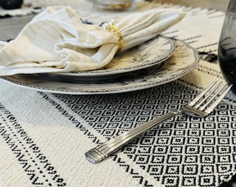 May include: A dining table setting with a cream and black patterned table runner, plates, a folded napkin, and a silver fork. A gold napkin ring and a glass vase with decorative branches are also visible.