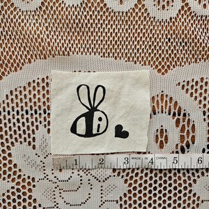 May include: A small, square fabric patch with a black bee illustration and a heart. The bee has a simple design with a body and antennae. A ruler shows the patch is approximately 3 inches wide.