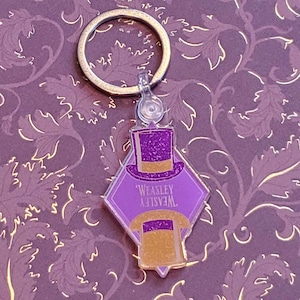 Puede incluir: Llavero morado y dorado con un sombrero de mago y el texto "WEASLEY" en un dije en forma de diamante.