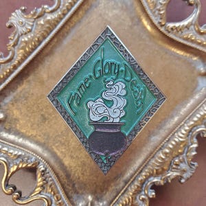 May include: A green diamond-shaped enamel pin with a cauldron and steam, surrounded by a mosaic-like border. The text "Fame, Glory, Death" is written around the cauldron.