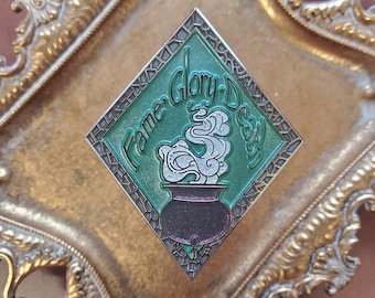 May include: A green diamond-shaped enamel pin with a cauldron and steam, surrounded by a mosaic-like border. The text "Fame, Glory, Death" is written around the cauldron.