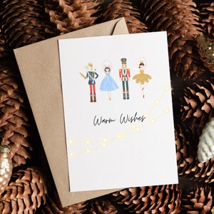 May include: A white card with a colorful illustration of a nutcracker, a ballerina, a princess, and a mouse. The illustration is surrounded by a string of white lights. The text "Warm Wishes" is written below the illustration.